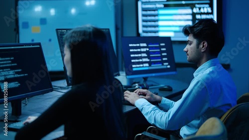 Focused young asian IT professionals work late typing on computer in dark, blue-lit cybersecurity control room. They actively analyze complex data networks and write software code on glowing monitors
