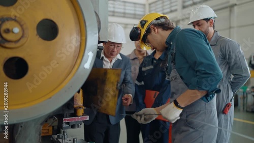Factory workers inspect heavy machinery with a supervisor. They wear protective gear and gloves, focused on their task. The team discusses plans using a tablet and clipboard