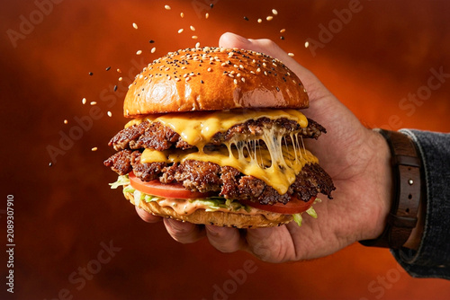 Quadro su tela Hand Holding Double Smash Burger with Melted Cheese on Dark Orange Background
A