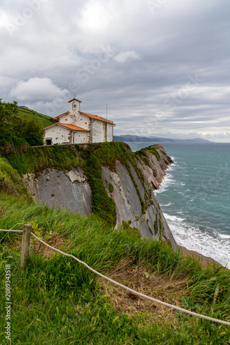 A small white chapel with an orange roof sits precariously on a steep cliff overlooking the Cantabrian Sea in Zarautz, Spain