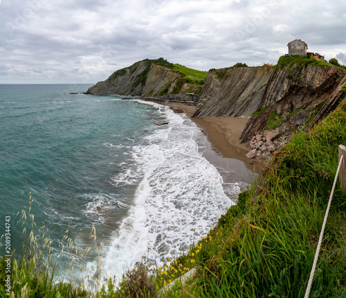 Dramatic geological formations of layered rock rise above a sandy cove with crashing waves on the Basque coast
