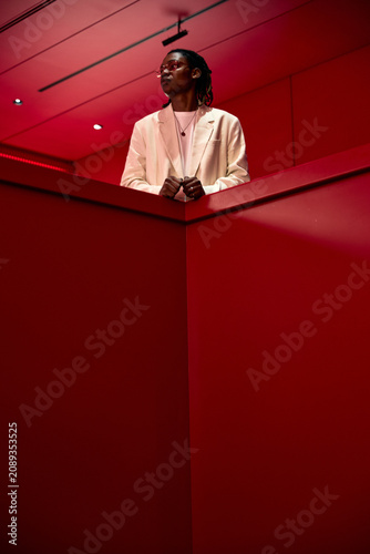 Vertical shot of young adult African American man standing behind geometric wall  looking away and posing in modern interior  conveying confidence and style. Ideal for fashion branding