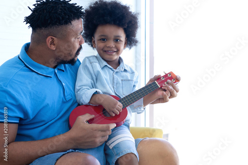 Son sat on his African father's lap, and they were both playing the ukulele together. boy looking at camera. Father's Day
