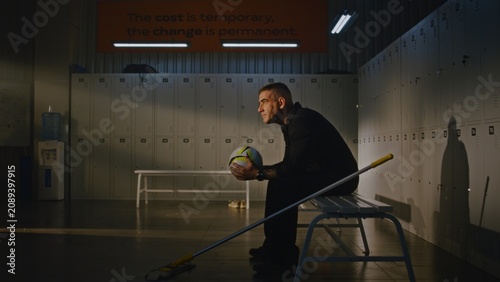 Tired Cleaner in Takes Break From Mopping Sports Locker Room. Man Sits on Bench, Picks Up Soccer Ball Left Behind by Team, and Holds it With Both Hands, Contemplates Youth With Sense of Nostalgia.