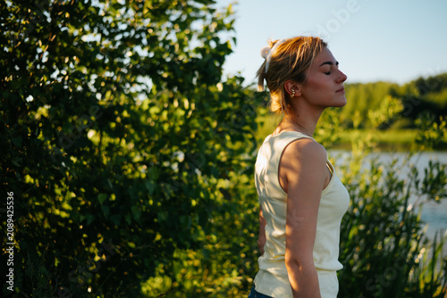 A woman stands still near a body of water, lost in thought while enjoying the warmth of the sun. The lush greenery surrounds her, creating a serene backdrop for this moment