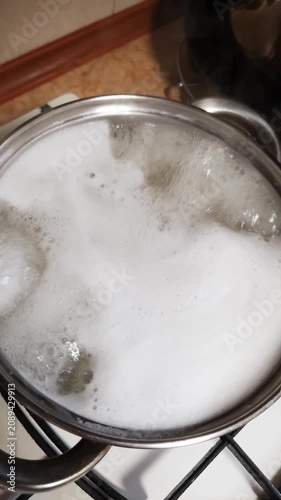 A top-down view of a stainless steel pot on a gas stove with hot liquid and thick white foam bubbling vigorously. Concept of cooking pasta, kitchen preparation, and boiling over.