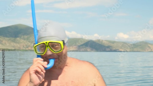 A funny overweight man puts a snorkel into his mouth while wearing swim goggles before diving. His comic appearance brings joy and laughter, funny fat man enjoying a beach vacation.