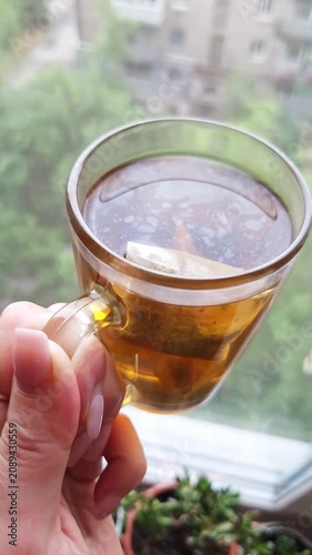A hand with pink manicured nails holds a clear glass mug filled with hot tea and a tea bag near a window. Concept of morning routine, relaxation, tea time, and healthy lifestyle.