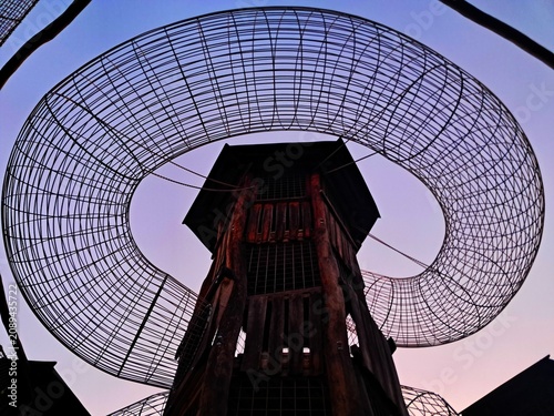 An abstract low angle view of a tall wooden play tower encircled by a massive metal wire climbing tunnel on a playground in Prague at twilight.