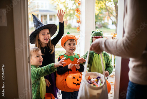 Children in witch, pumpkin and dinosaur costumes receiving halloween candy while trick or treating at a neighborhood front door.