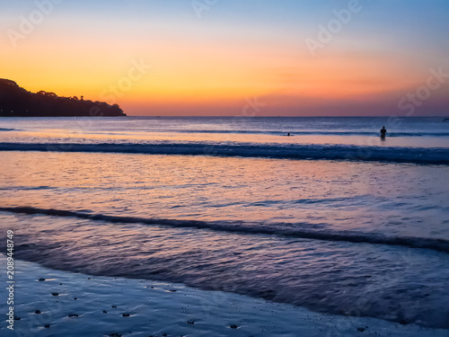 Tropical beach at twilight with orange and blue sunset sky, silhouetted headland and fishing boat on calm ocean