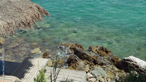 Waves crushing of beachfront wooden deck with sun loungers above rocky turquoise sea of Adriatic sea on Albanian RIviera