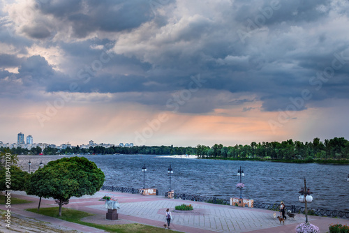 Dramatic sunset after a thunderstorm over Obolon Embankment in Kyiv, Ukraine. Golden sunbeams break through storm clouds, reflecting across the Dnipro River and city skyline.
