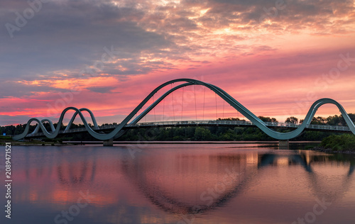 Sunrise over the pedestrian bridge across the Dnipro River in Kyiv, Ukraine. Golden sunlight breaks through pink clouds, reflects on calm water, and slowly rises above the city skyline.