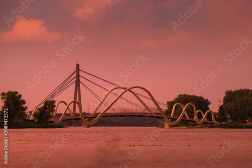 Golden sunset light after a powerful thunderstorm illuminates Wave Bridge and North Bridge over the Dnipro River in Kyiv, Ukraine, creating a dramatic cityscape with glowing reflections.