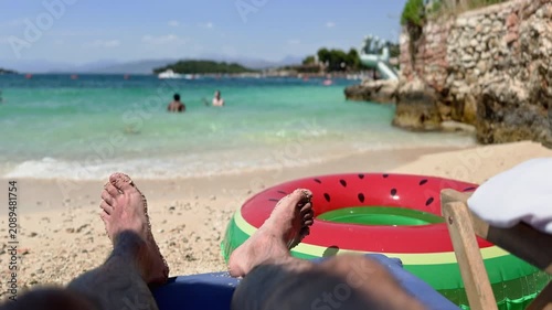 First-person view of a man relaxing on a sunny sandy beach sunbed near crystal-clear turquoise water on the Albanian coast