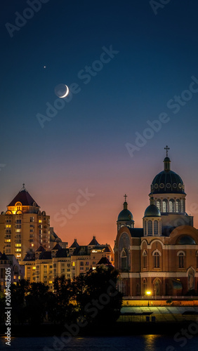 Night view of Obolon Embankment in Kyiv, Ukraine, with a thin crescent Moon above illuminated church domes and city skyline reflected in the peaceful Dnipro River at blue hour.