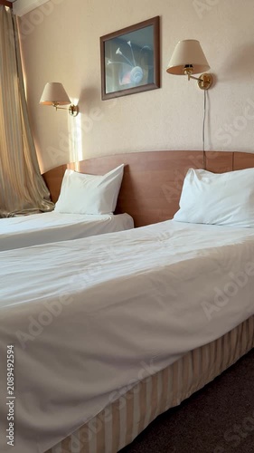 Bright economy hotel room twin beds, white linen and plump pillows on neatly made beds, wooden headboard with brass wall sconces