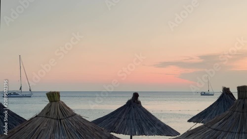 Straw beach umbrellas at sunset with sailboats on calm sea