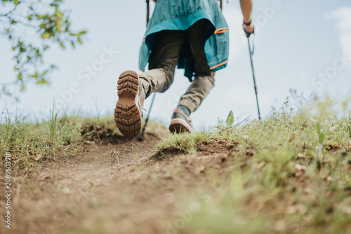 Fotografie A hiker climbs a dirt trail using trekking poles, shown from a low rear angle