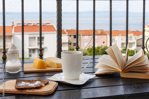 Breakfast table on a balcony in Makarska, Croatia, with coffee cup, open book, bread with butter, and Adriatic Sea view
