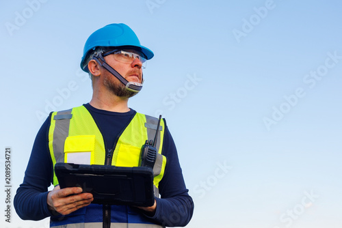 Fotografie Inspector and technician installer wearing a blue hard hat, safety glasses, and reflective vest, holding a digital tablet during an inspection work at an outdoor construction site
