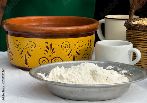 White wheat flour in a vintage metal plate on a white tablecloth, with a traditional decorated yellow Cretan clay basin and ceramic mugs in the background. Authentic food still life.