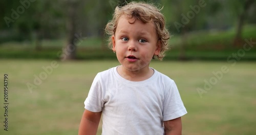 Wide-eyed, thoughtful baby boy stands in park at evening. He briefly responds, then glances away, lost in curiosity. Slow-motion portrait captures his innocent expression and gentle emotion
