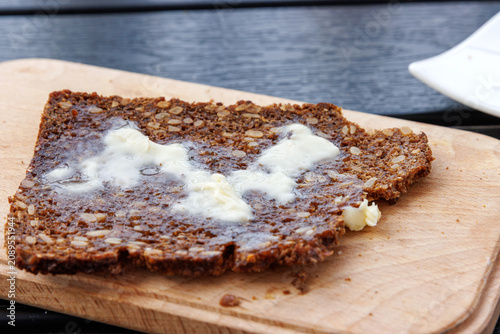 Closeup of a textured slice of sourdough wholegrain rye bread with nuts and grains, topped with melting butter, on a wooden board.