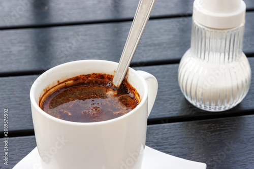 Closeup of black coffee in a white cup with spoon on a dark wooden table, with a sugar dispenser in the background. 

