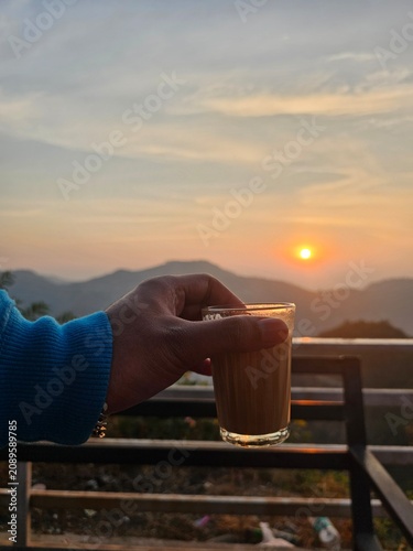 A hand holding a glass of tea against a backdrop of a serene sunset, with mountains in the distance, evoking a tranquil and peaceful evening atmosphere.