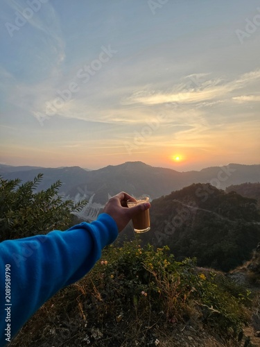 A hand holding a glass of tea against a stunning sunset backdrop, showcasing the beauty of nature and tranquility in the hills.