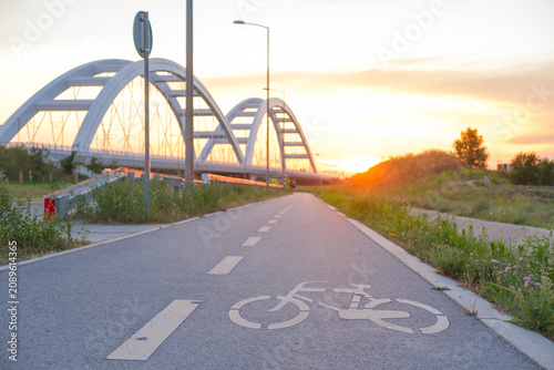Bicycle path along a waterway at sunset, curved bridge arches in the background, paved lane with bike marks, peaceful outdoor scene.