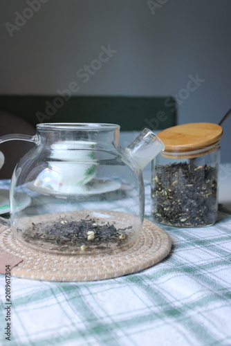Glass teapot filled with loose tea leaves placed on a woven mat next to a jar of dried herbs on a checkered tablecloth in a cozy indoor space