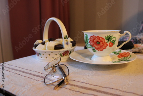 Floral-patterned porcelain tea cup sits next to a basket of sweets on a table, with reading glasses resting nearby in a cozy indoor environment