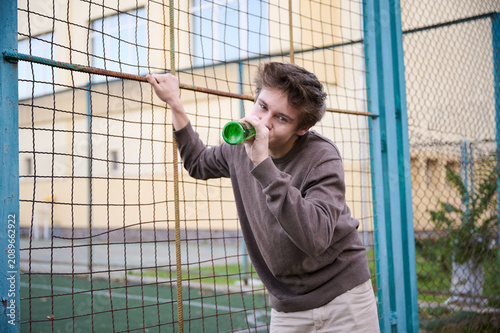 Картината върху платно Teen boy drinks from bottle beside sports area fence