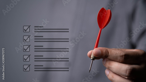 Close-up of a hand holding a red dart next to a checklist interface, symbolizing goal achievement, business planning, task management, strategic focus, project execution, and performance improvement.