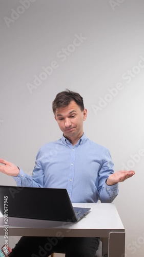 Vertical video man wearing a blue shirt sitting at a desk with a laptop. Looking confused and shrugging with open hands. Expressing uncertainty. Doubt