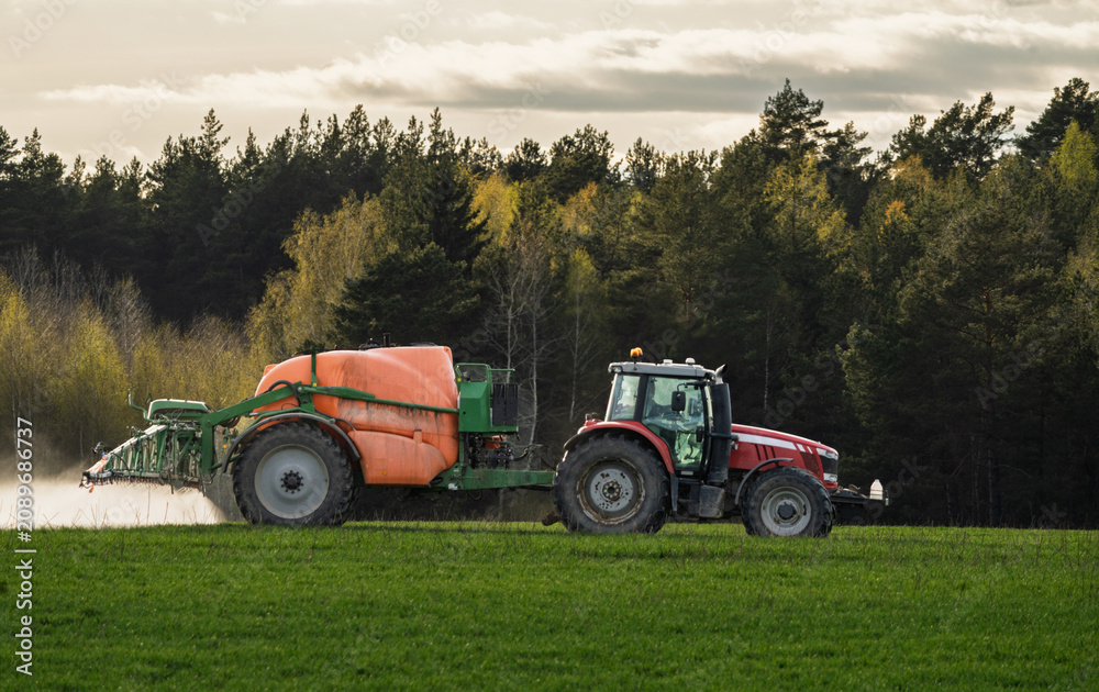Obraz premium Tractor With Mounted Sprayer Applying Fertilizer On Green Field Near Forest At Dusk