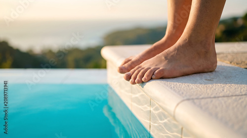 Feet dangling over edge of swimming pool at sunset with soft light reflecting on water surface during peaceful outdoor vacation evening.