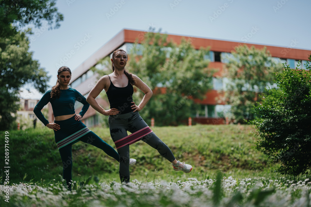 Obraz premium Two women perform standing leg exercises with resistance bands for fitness and strength. The pair train together outdoors on grass near an urban building, practicing balance and mobility.