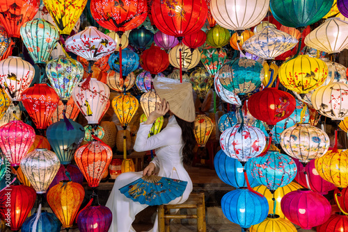 An Asian woman in an ao dai is sitting and admiring the colorful lanterns in the city of Hoi An, Vietnam.
