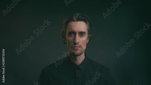 Moody studio portrait of solitary adult man in black shirt, subtle smile and raised eyebrows, facing camera against dark blurred background with soft dramatic lighting
