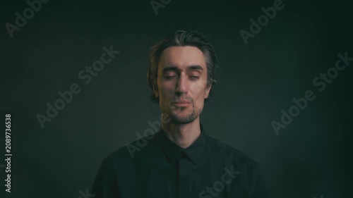 Close up studio portrait of young adult man with dark hair in black button up shirt, changing expressions from neutral to slight smile to serious, dark background