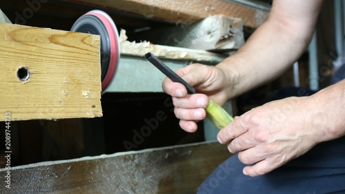 A craftsman sharpens a chisel in a woodworking shop. The man's hands carefully guide the tool's metal blade to the rotating grinding wheel of a sharpening device near a wooden workbench.