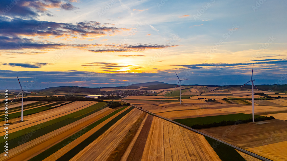 Fototapeta premium Wind turbines over rural fields at sunset. Drone view with village, hills, golden crops, layered clouds.