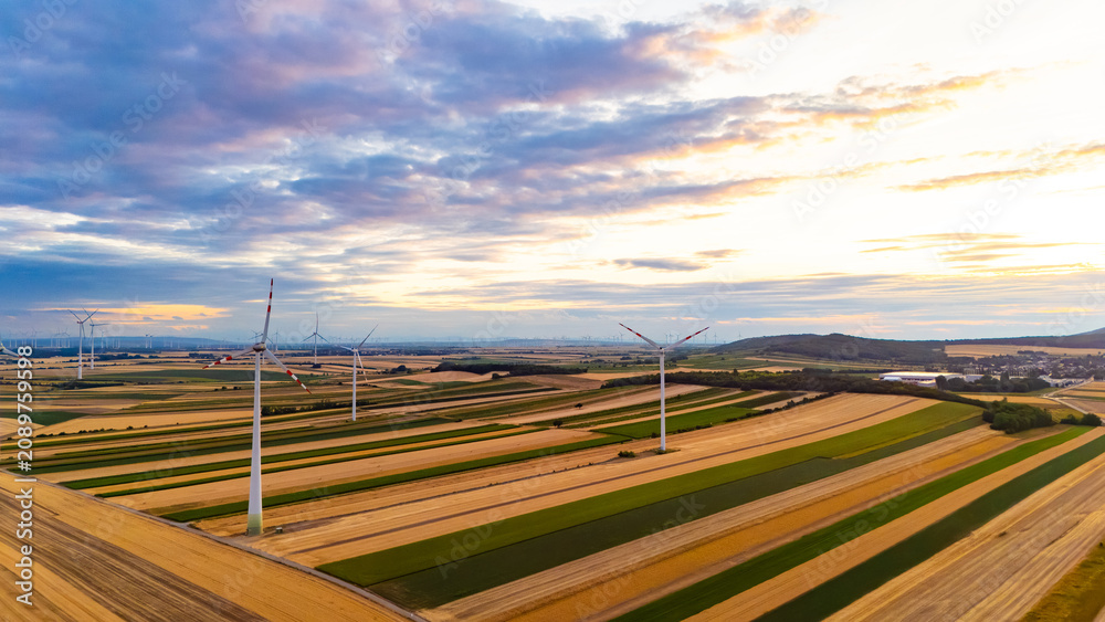 Fototapeta premium Wind turbines stand tall in green and yellow fields during sunset. Wind turbines spin in a landscape of fields at sunset. The sky is bright with colors.