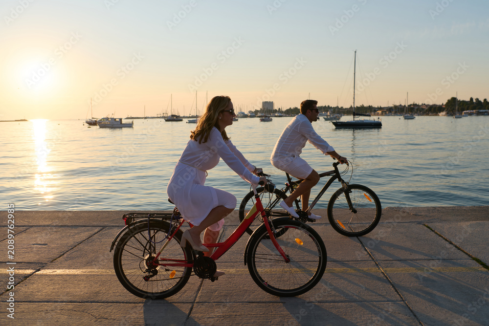 Fototapeta premium Couple Cycling Along Waterfront At Sunset Near Sailboats On Summer Evening