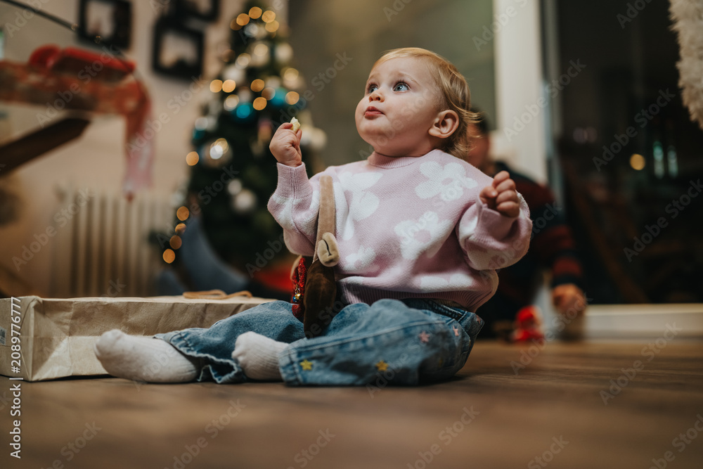 Fototapeta premium A toddler girl sits on the floor in a pink floral sweater, looking up with curiosity. A cozy home scene shows a Christmas tree and gifts nearby, capturing a warm family moment.