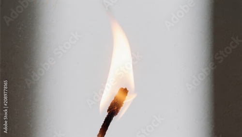 A close-up image of a burning matchstick with a bright flame against a plain background.